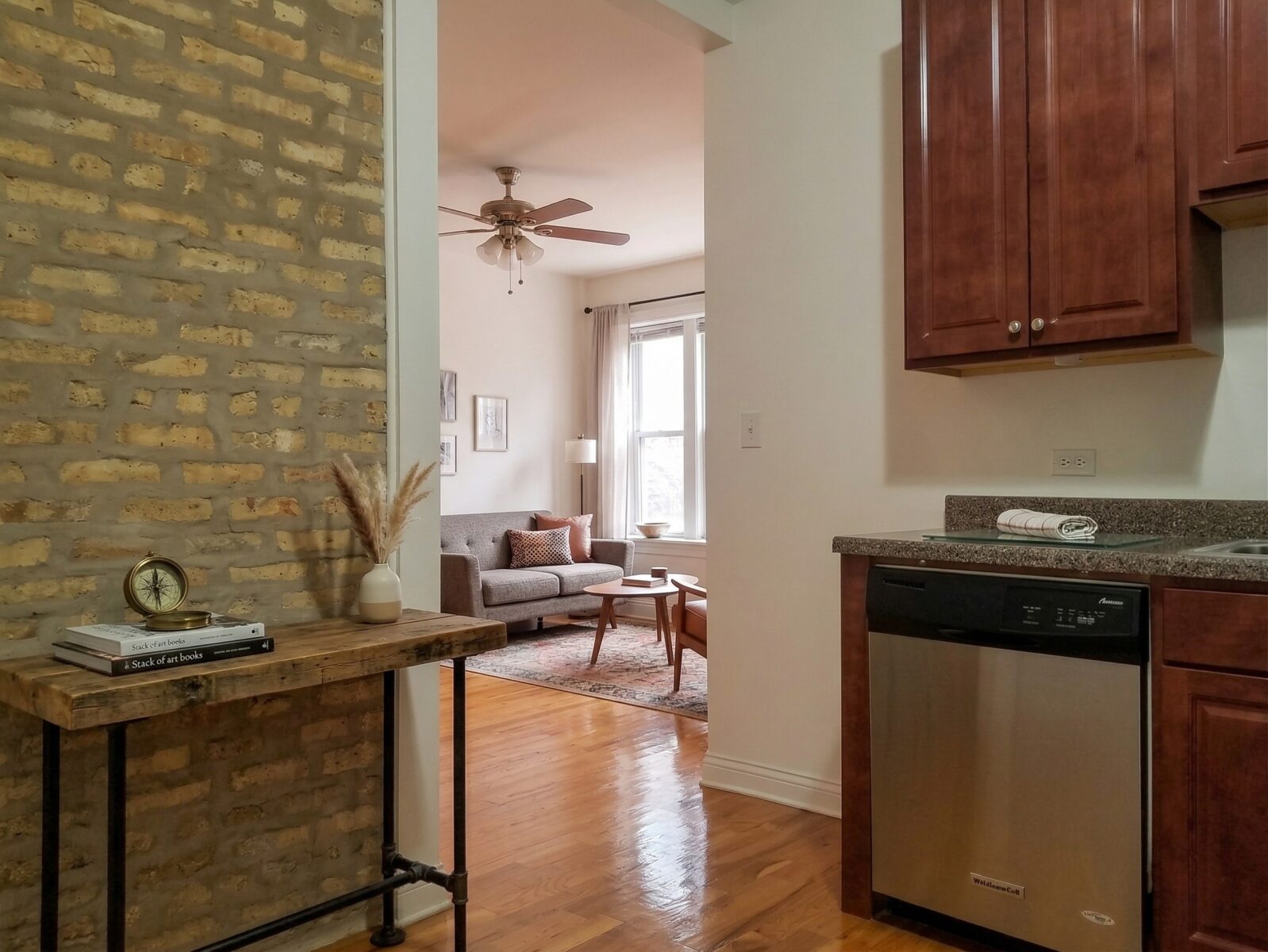Living room featuring an exposed brick wall, hardwood floors, and a view into a modern kitchen.