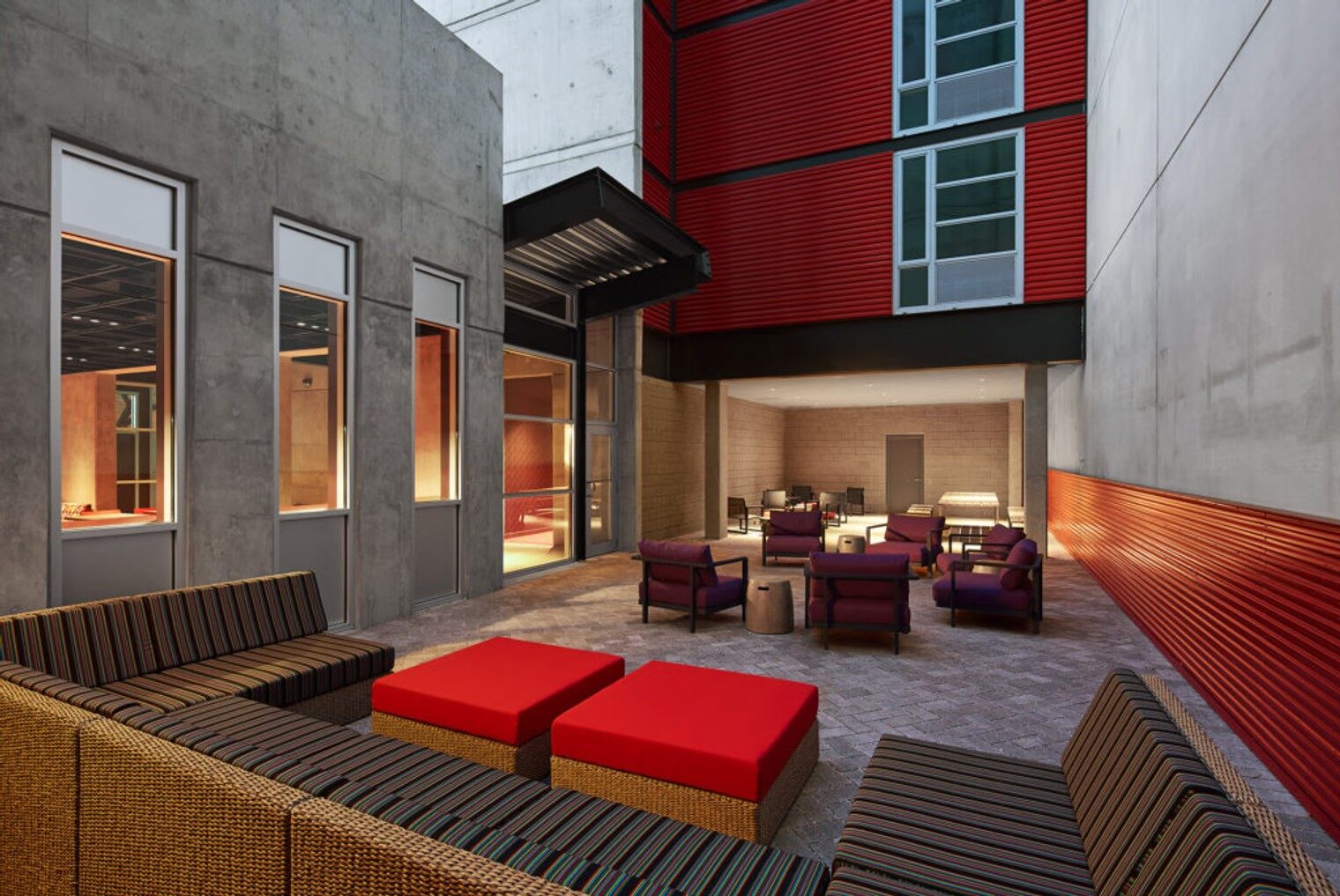 Outdoor courtyard with concrete walls, red facade panels, paved flooring, and covered seating areas.