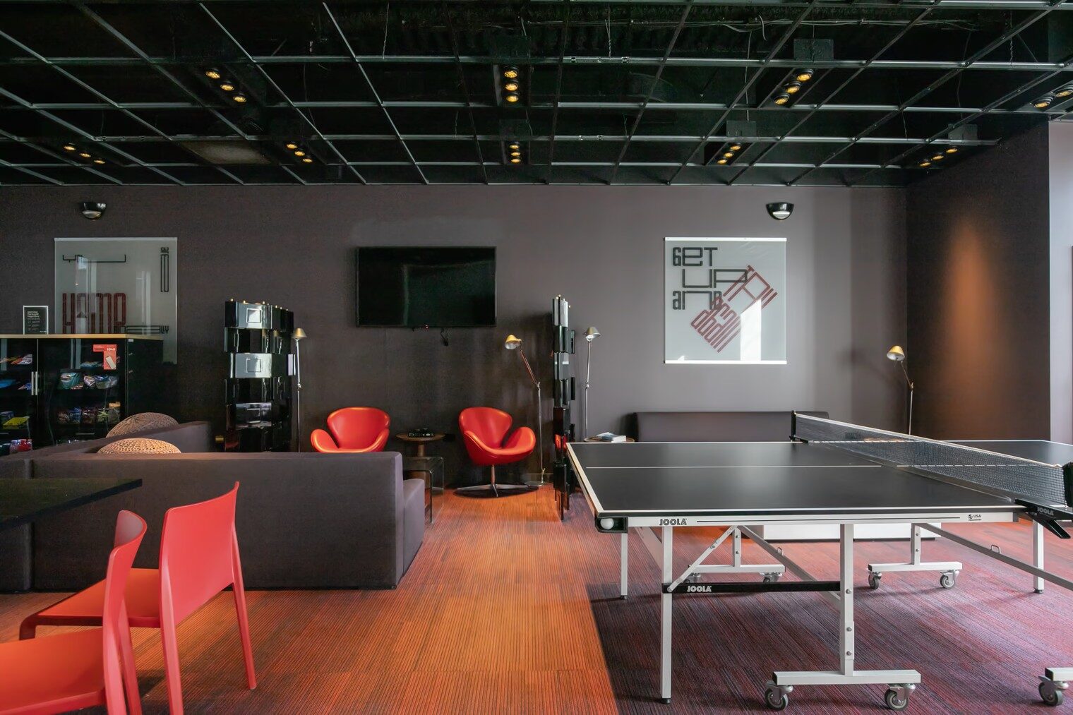 Resident lounge with ping-pong table, seating areas, wall-mounted television, and exposed ceiling.