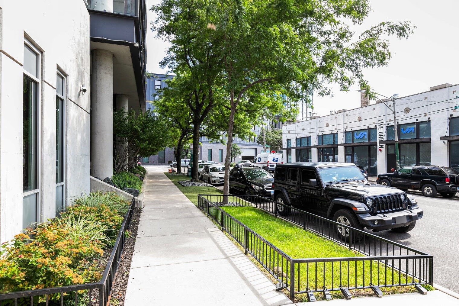 Building exterior walkway with landscaped planting beds, mature trees, and metal fencing.