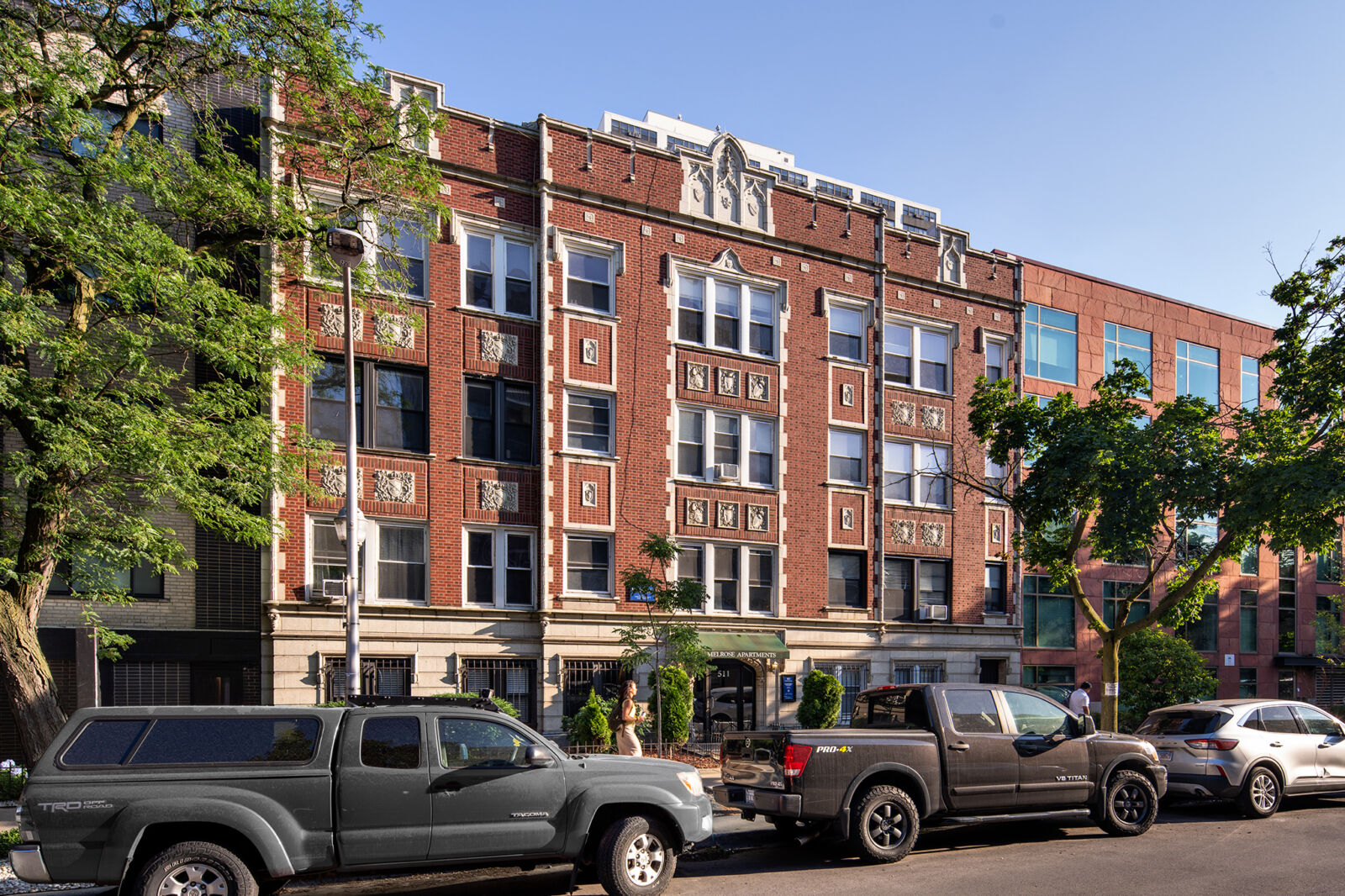 Exterior of a vintage brick apartment building on a sunny day with cars parked along the street.
