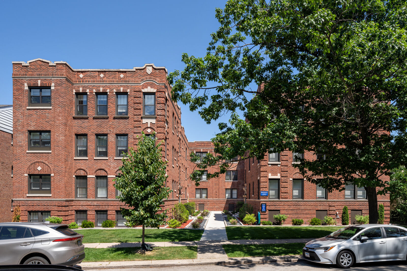 Three-story red brick apartment building with courtyard entrance and landscaped walkway, located ...